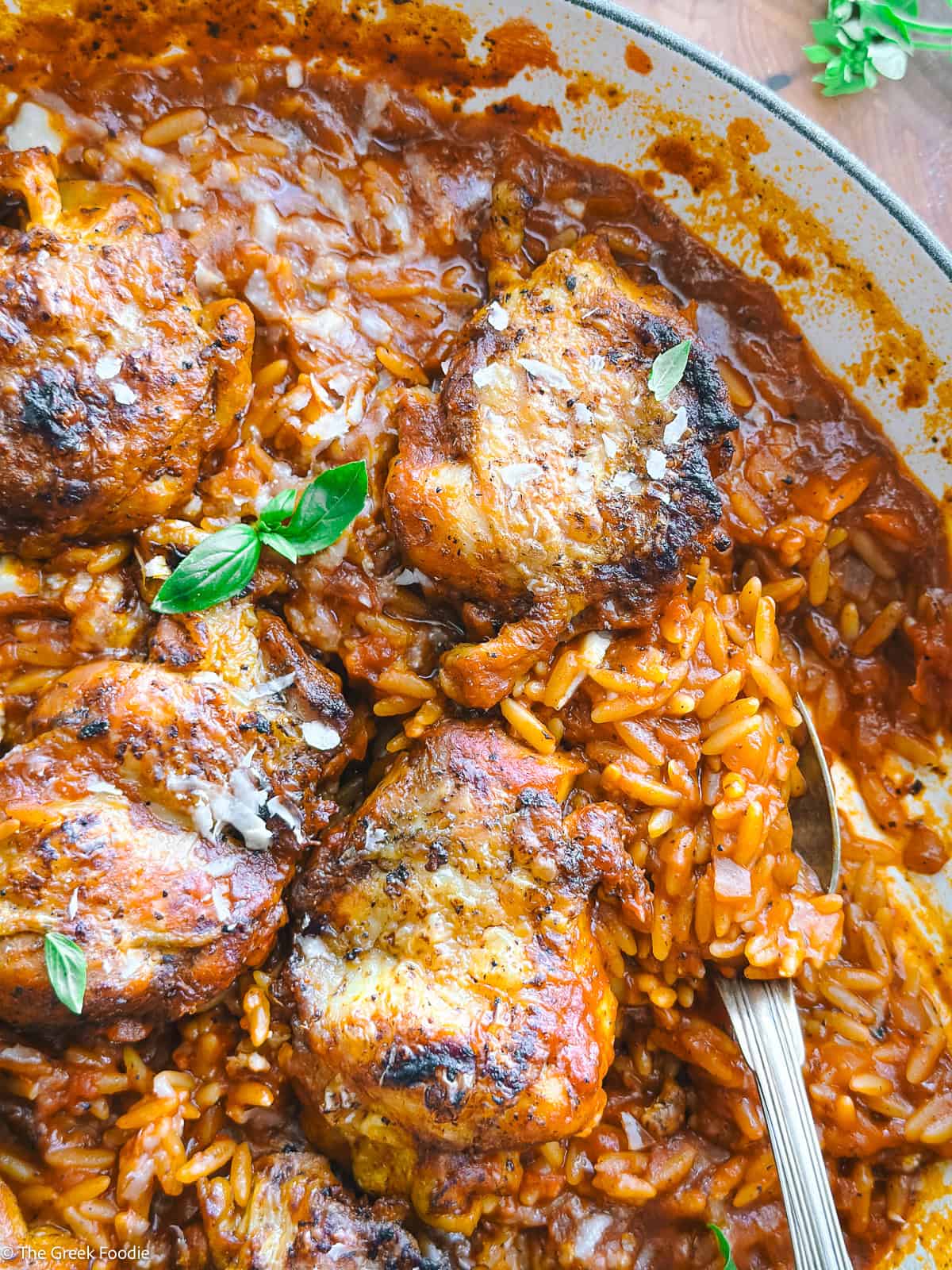 Side view of a pan of chicken youvetsi with orzo (kritharaki) in tomato sauce, topped with herb leaves.