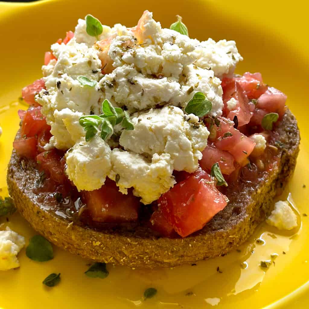 A rusk with feta cheese and chopped tomatoes on a yellow plate.