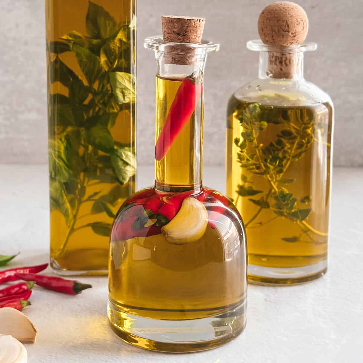 Three glass bottles filled with olive oil, various herbs and garlic cloves on a white table.