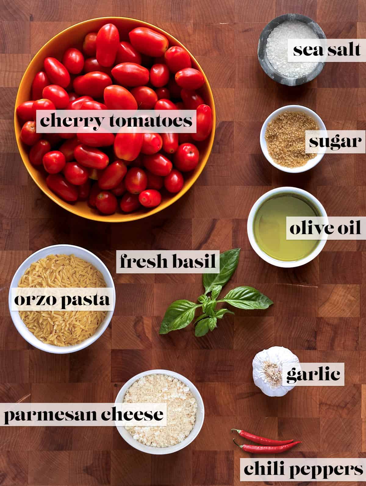 Tomatoes, raw orzo, sea salt, sugar, fresh basil, olive oil chili peppers and parmesan cheese in separate containers on a table.