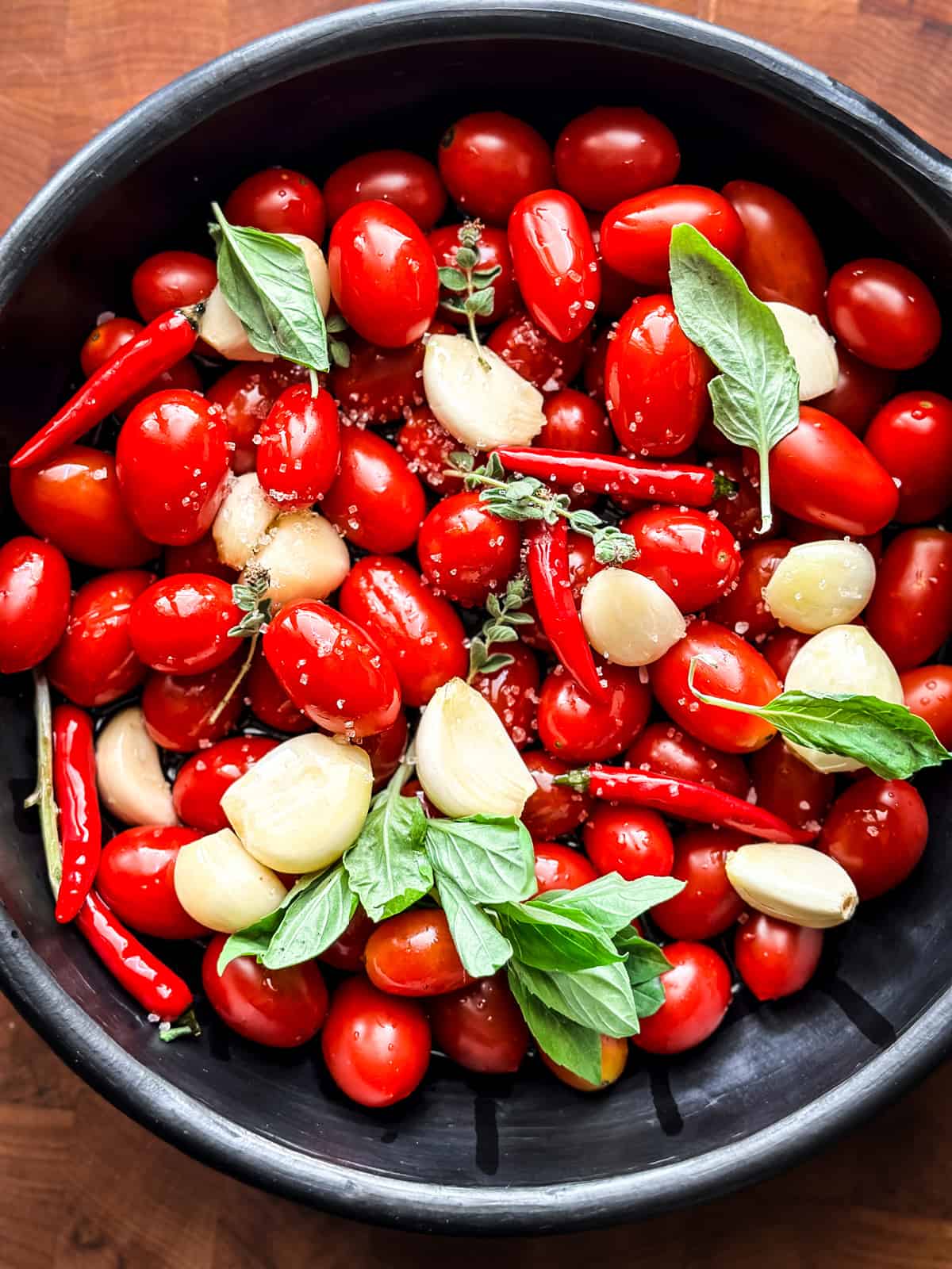 A round black baking pan with cherry tomatoes, fresh basil and garlic cloves in olive oil on a wooden table.