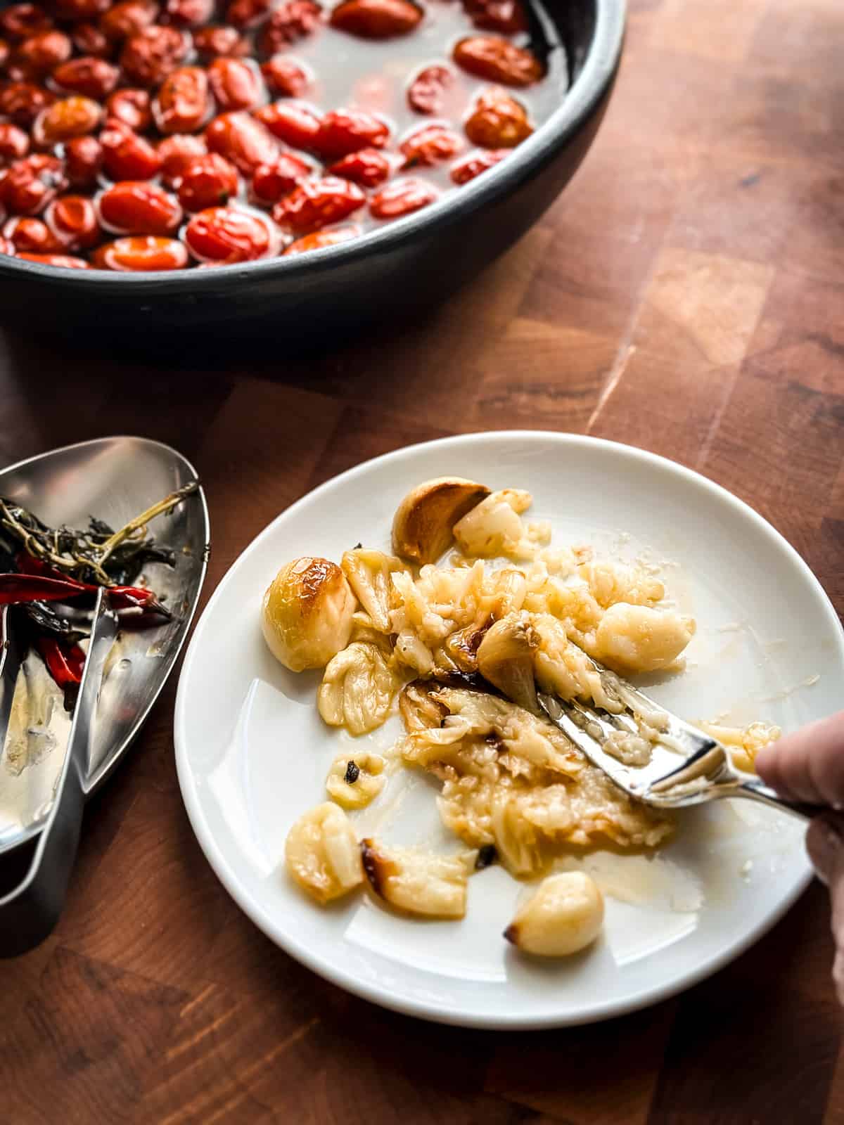 A hand is crushing roasted garlic cloves on a late with a fork. Partial view of a baking pan with tomatoes and a small plate with chili peppers and herbs.