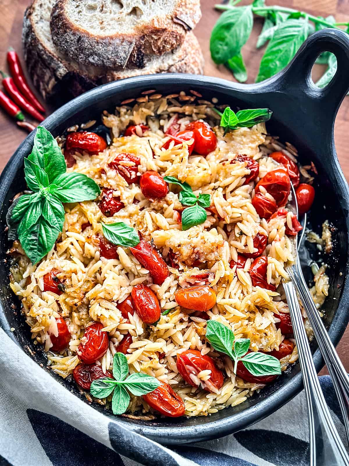 A round baking pan with orzo pasta, tomatoes, fresh basil leaves and parmesan with serving utensils on a wooden table.