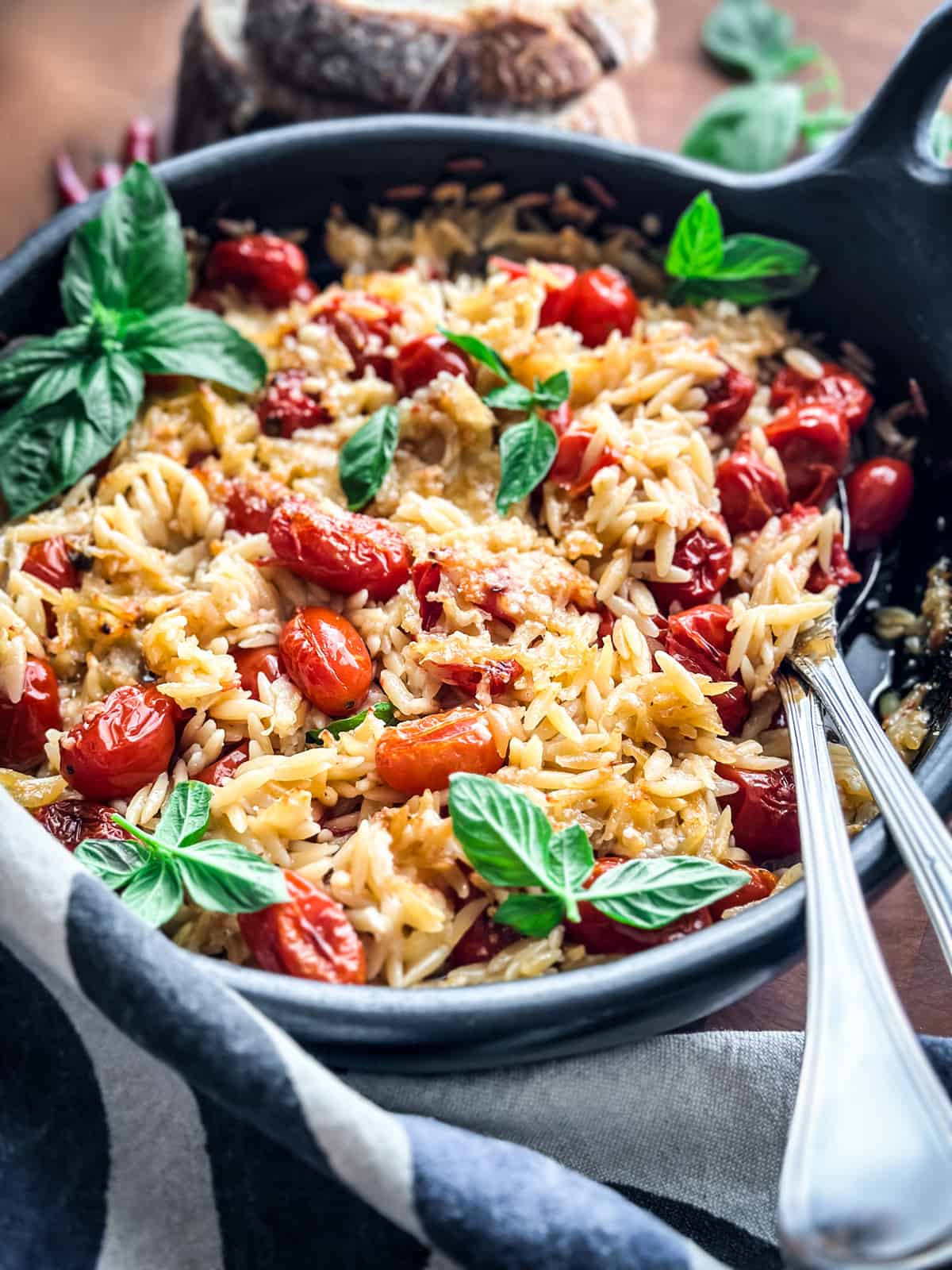 A round baking pan with orzo pasta, tomatoes, fresh basil leaves and parmesan with serving utensils on a wooden table.