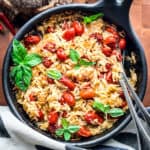A round baking pan with orzo pasta, tomatoes, fresh basil leaves and parmesan with serving utensils on a wooden table.