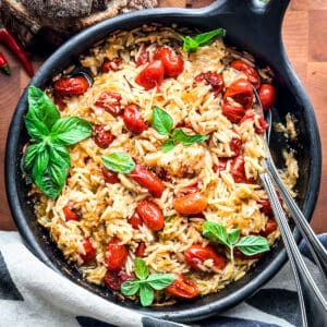 A round baking pan with orzo pasta, tomatoes, fresh basil leaves and parmesan with serving utensils on a wooden table.