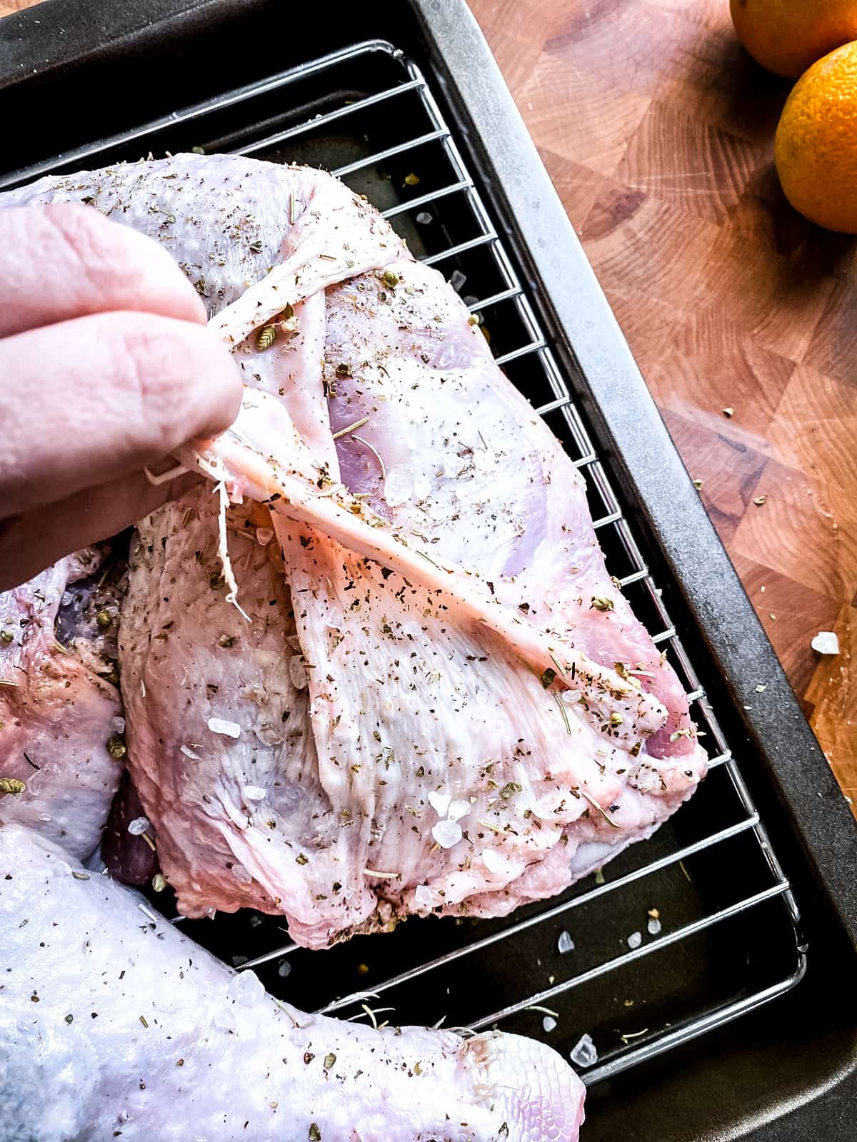 A hand lifting the skin from a turkey thigh with coarse salt and dried herbs on a wire rack.
