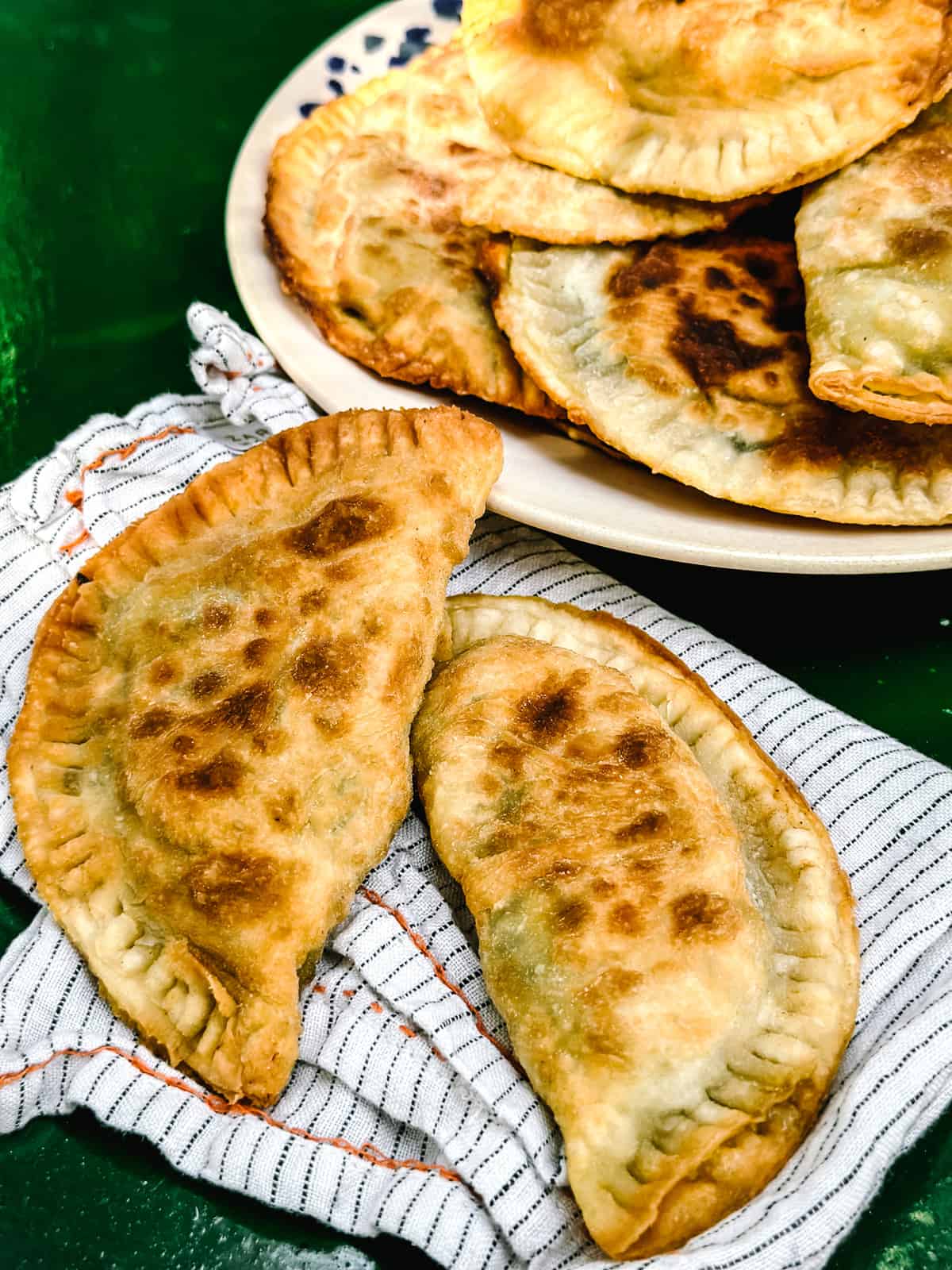 Kalitsounia, Cretan hand pies from Chania in a plate and two hand pies on a cloth napkin all on a dark green table.