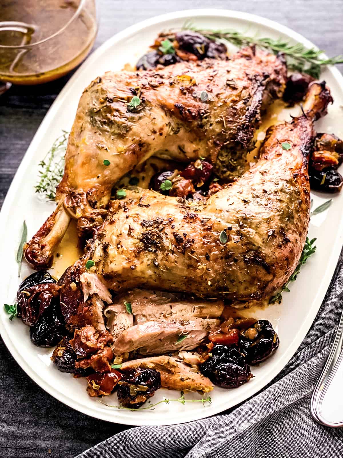 Two roasted turkey thighs with drumsticks on a platter with herbs and dry fruit, and gravy. Serving utensils are on one side and a bowl with gravy on the other.