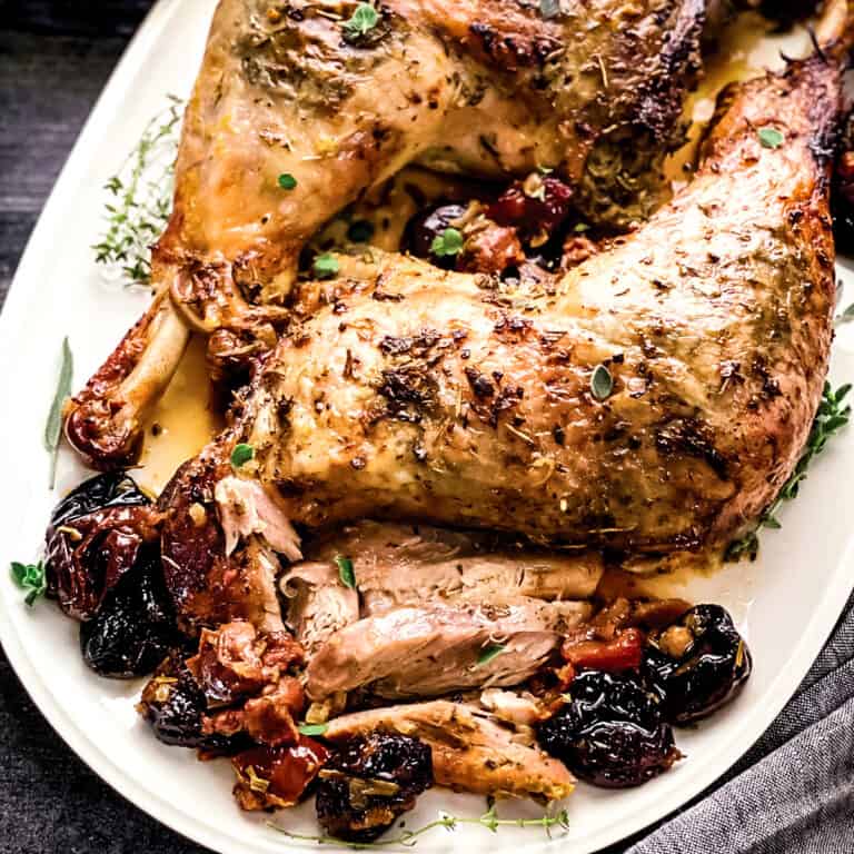 Two roasted turkey thighs with drumsticks on a platter with herbs and dry fruit, and gravy.