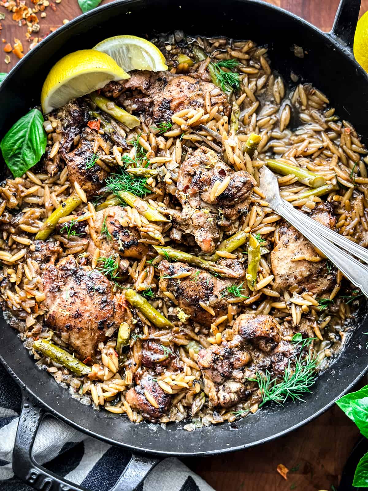 A skillet with Za'atar Lemon Chicken Orzo, two lemon wedges and serving utensils, and a cloth napkin on a table.