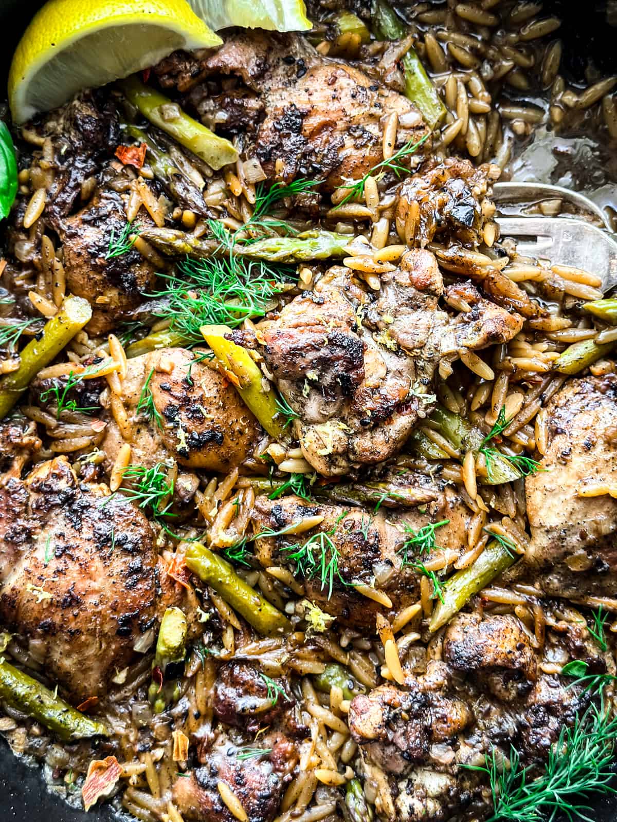 Close up of skillet with Za'atar Lemon Chicken Orzo, two lemon wedges and serving utensils.