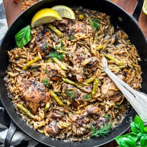 A skillet with Za'atar Lemon Chicken Orzo, two lemon wedges and serving utensils, and a cloth napkin on a table.