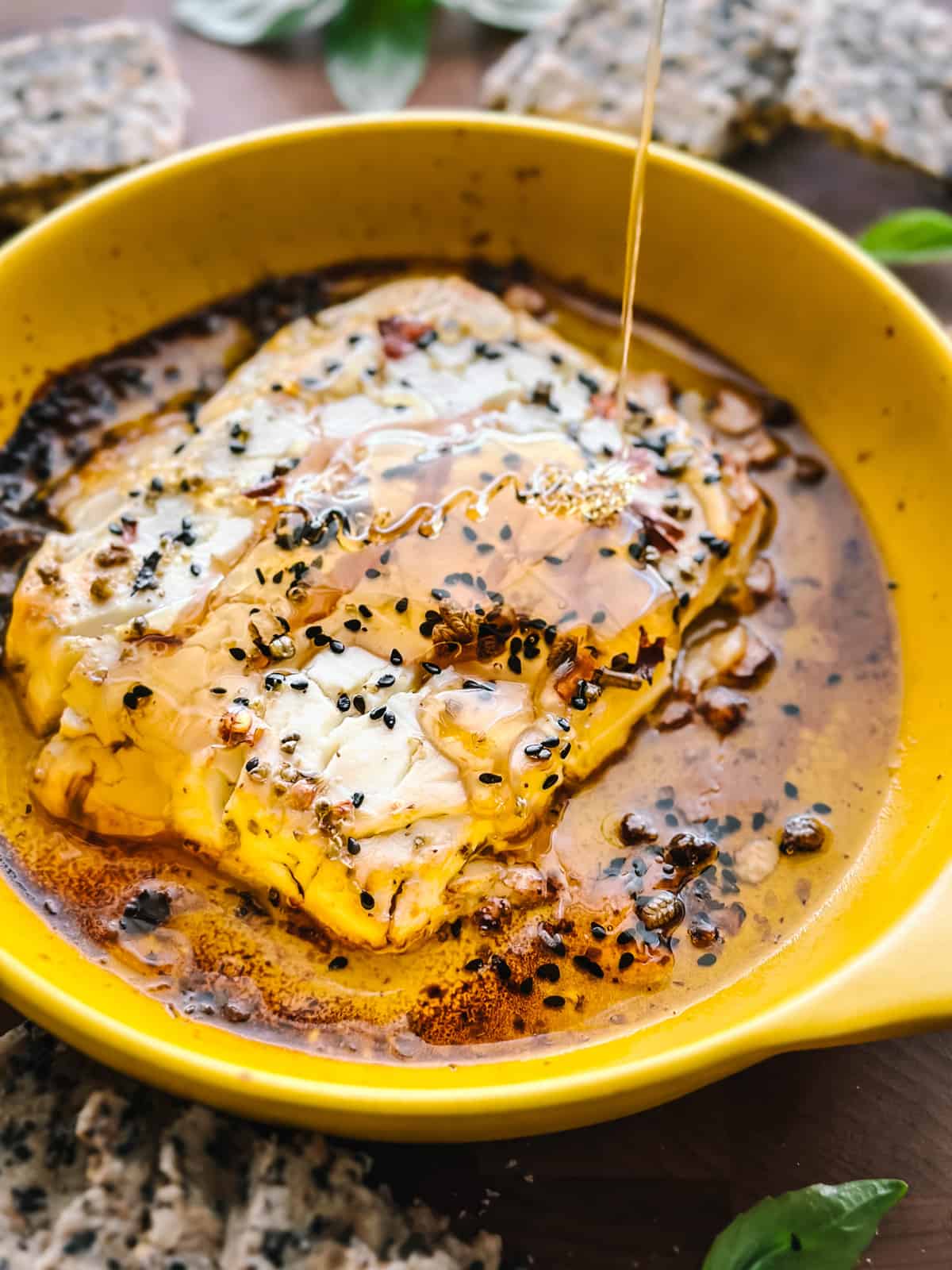 Baked feta cheese in a yellow round baking dish with seasonings and olive oil and honey dripping from above.