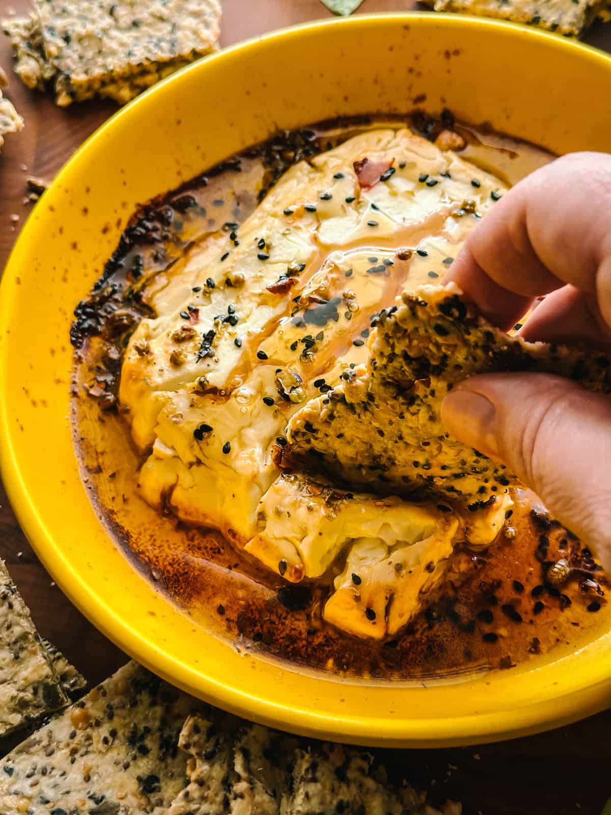 A hand holding a cracker scooping up baked feta cheese in a yellow round baking dish with seasonings and honey on top.