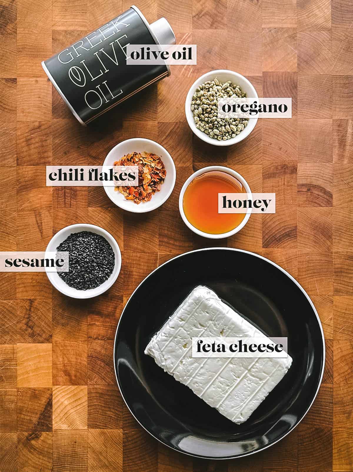 A bottle with olive oil, small conatiners with black sesame seeds, oregano buds, honey and chili flakes and a plate with feta cheese on a table. Overhead shot. 