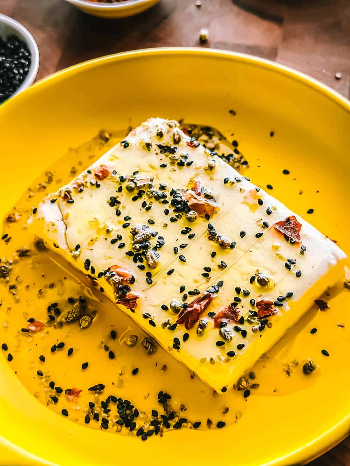 A block of feta cheese in a yellow round baking dish with seasonings and olive oil.