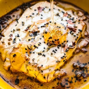 Baked feta cheese in a yellow round baking dish with seasonings and olive oil and honey dripping from above.