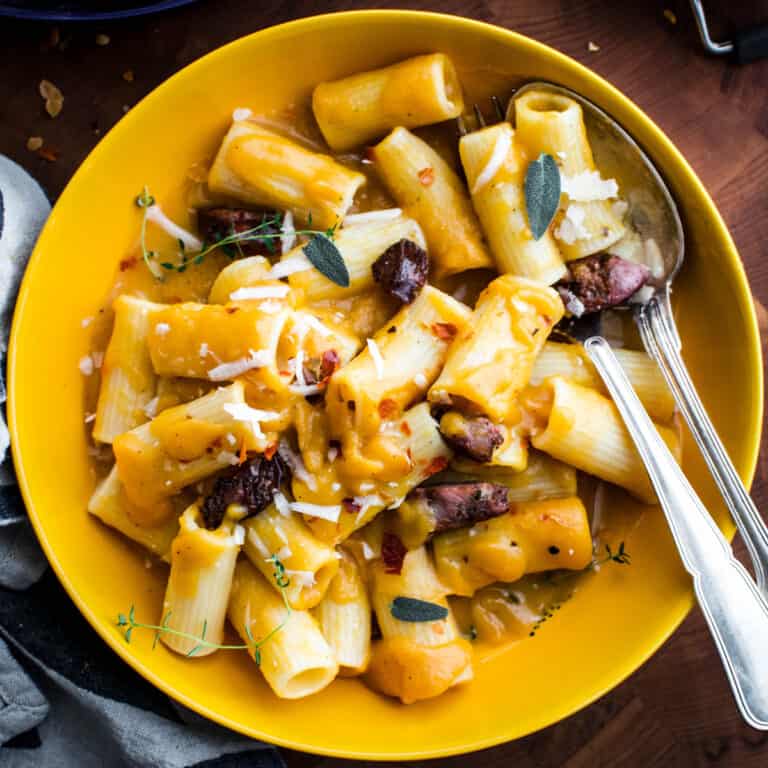 A plate with butternut squash pasta with sausages, a cloth napkin and utensils.