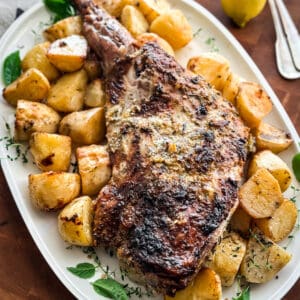 A Greek roasted leg of lamb with potatoes in a serving platter on a table with lemon halves around and herbs.