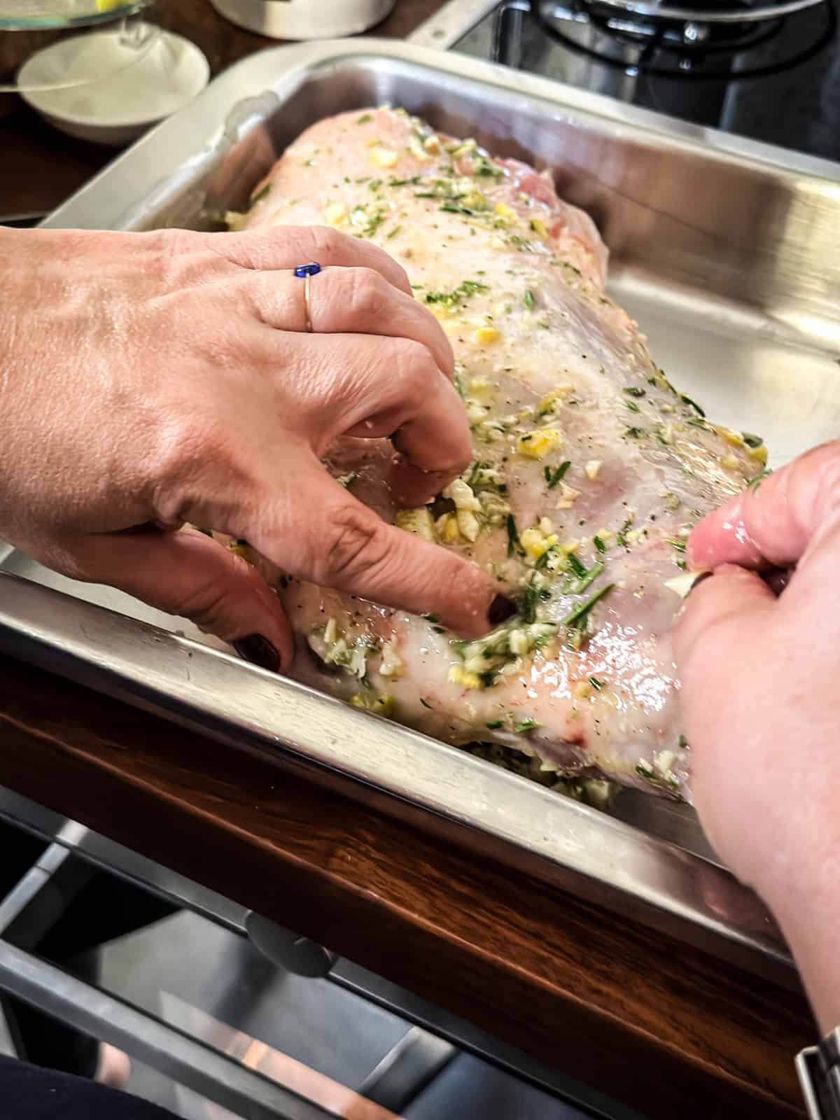 Hands rubbing marinade on a leg of lamb in a roasting pan on a kitchen counter.