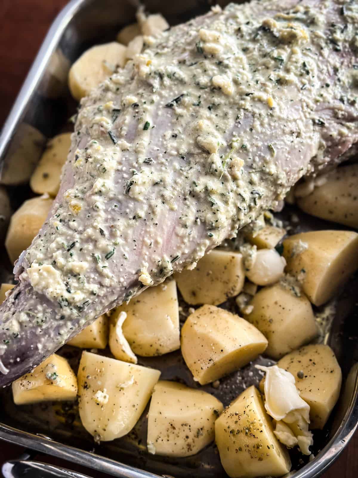 A leg of lamb with marinade on top of potatoes in a roasting pan.