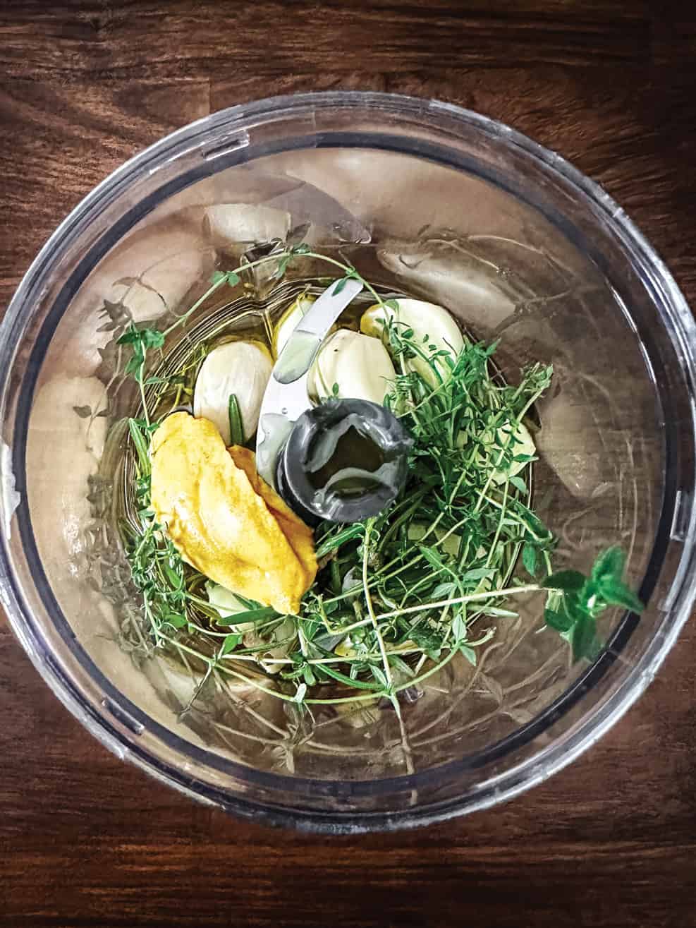 A food processor bowl with garlic, thyme sprigs, and dijon mustard.