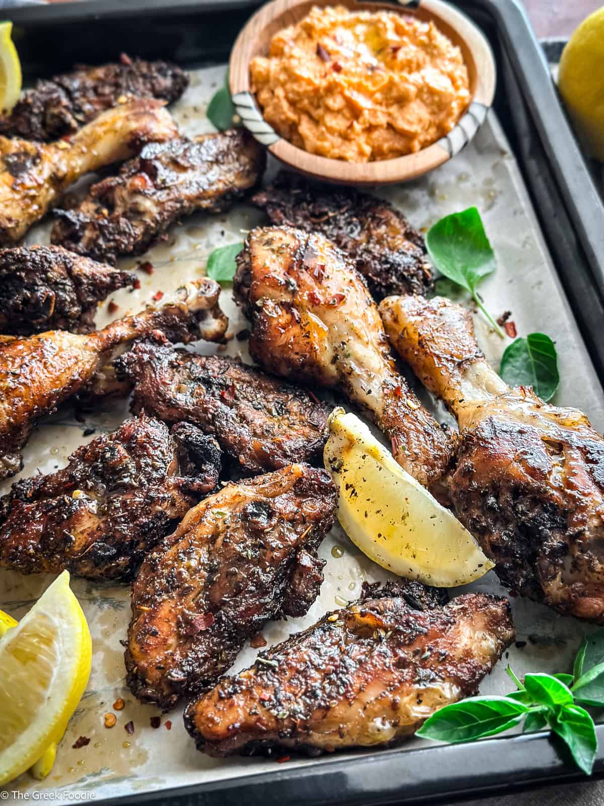 Greek chicken chops on a baking sheet with lemon wedges and whipped feta dip