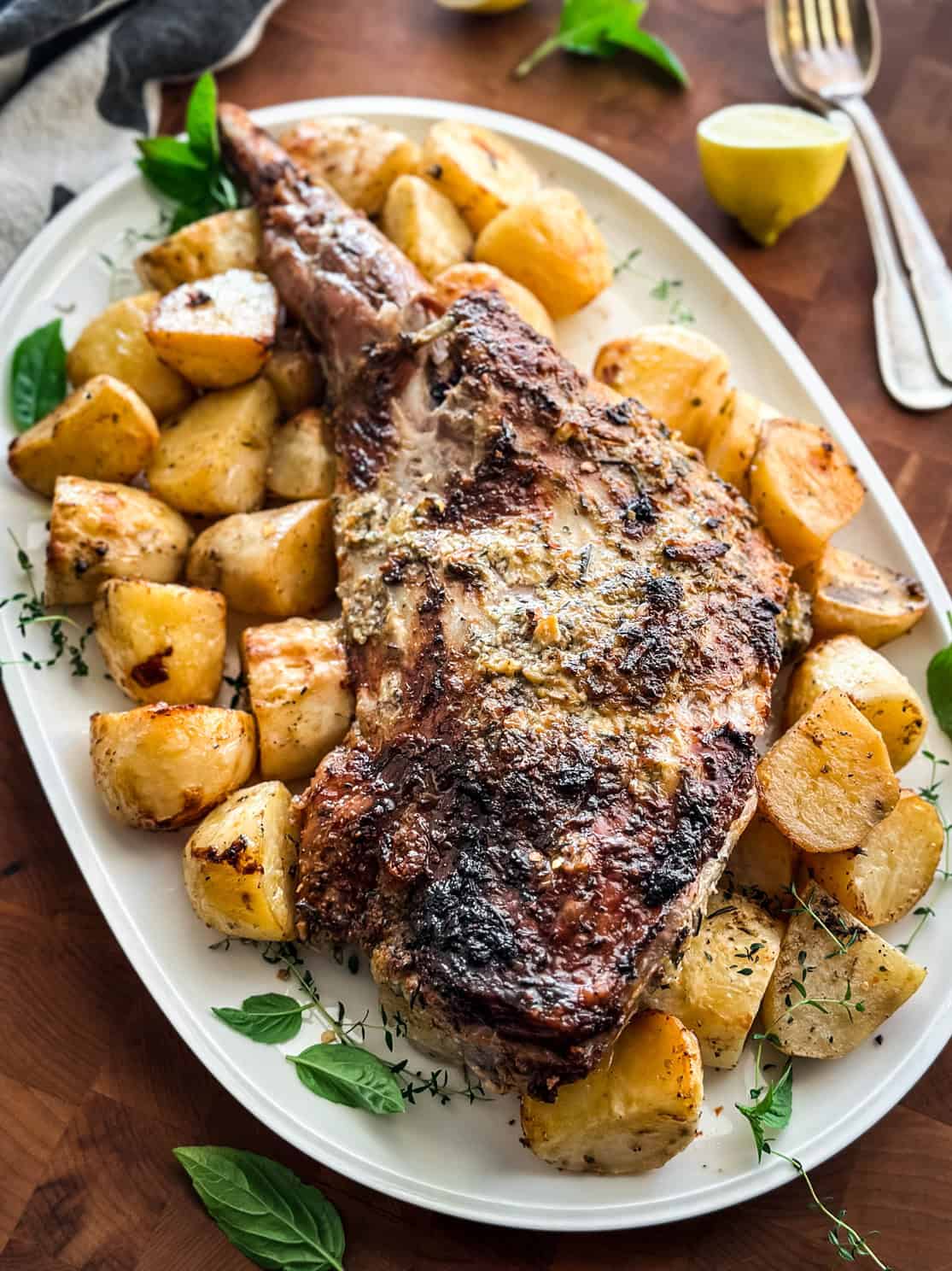 A Greek leg of lamb recipe slow roasted with potatoes on a serving platter with lemon halves and fresh herbs.