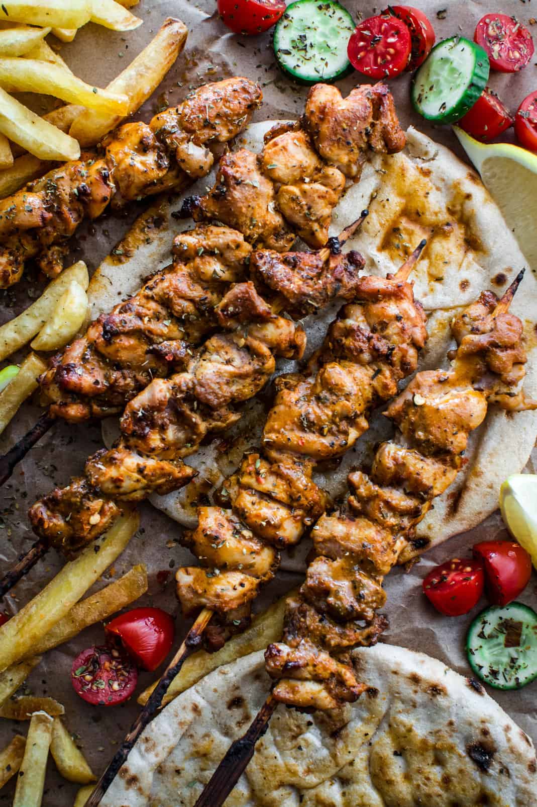Chicken thigh skewers on pita, cherry tomatoes, sliced cucumber, fries and lemon wedges on parchment paper.