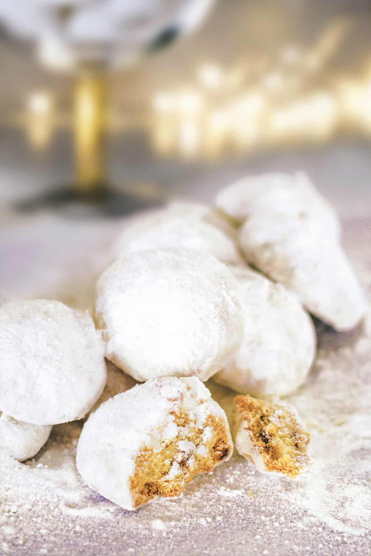 Kourabiedes- Greek Christmas butter cookies covered in powdered sugar on a table, a couple broken in half. At the back fading Christmas lights.