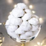 A glass bowl with Christmas butter cookies with two cookies on the table infant and Christmas lights at the back.