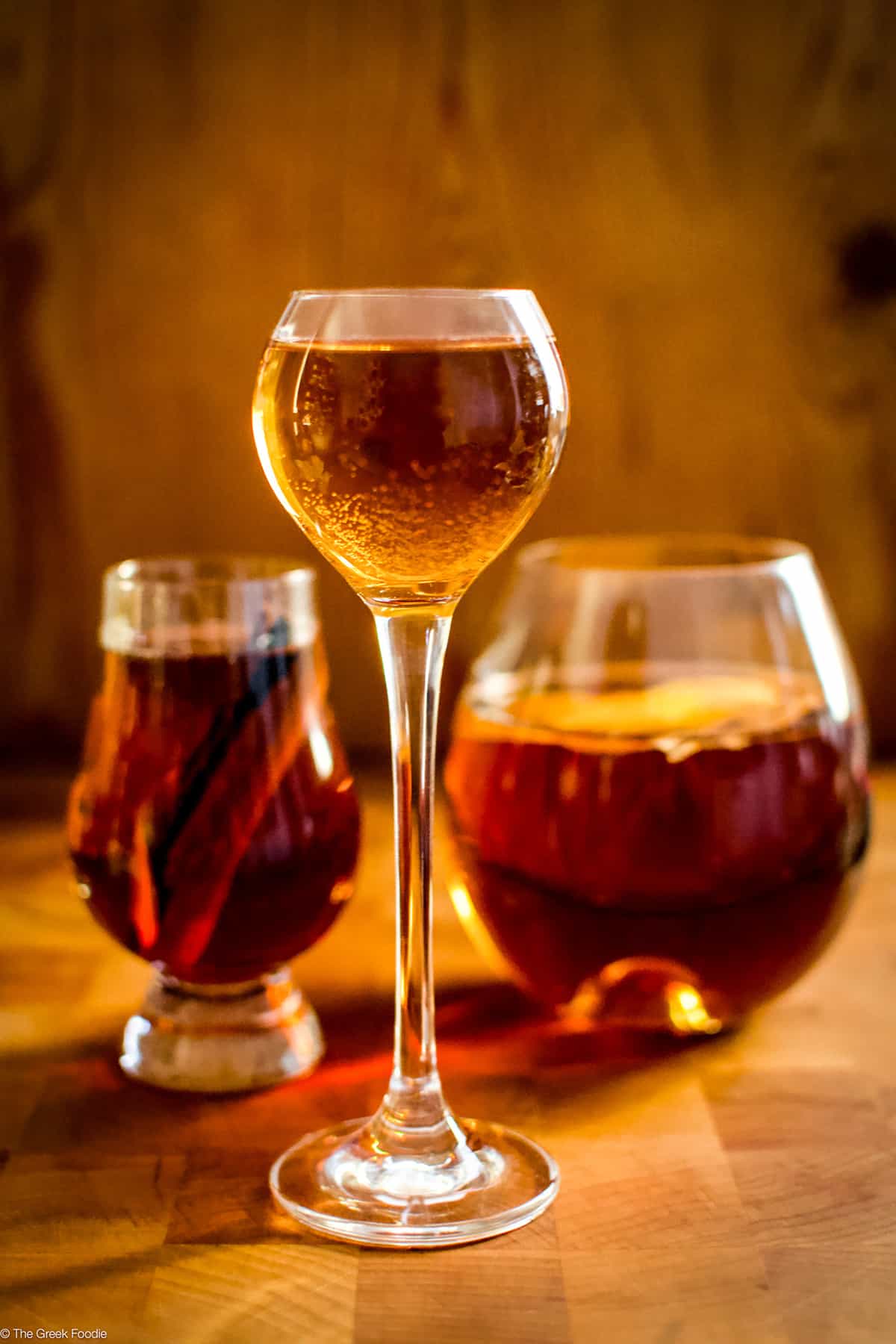 Three glasses, a tall small one, a big round and a medium glass with rakomelo, raki drink with honey in a wood environment.