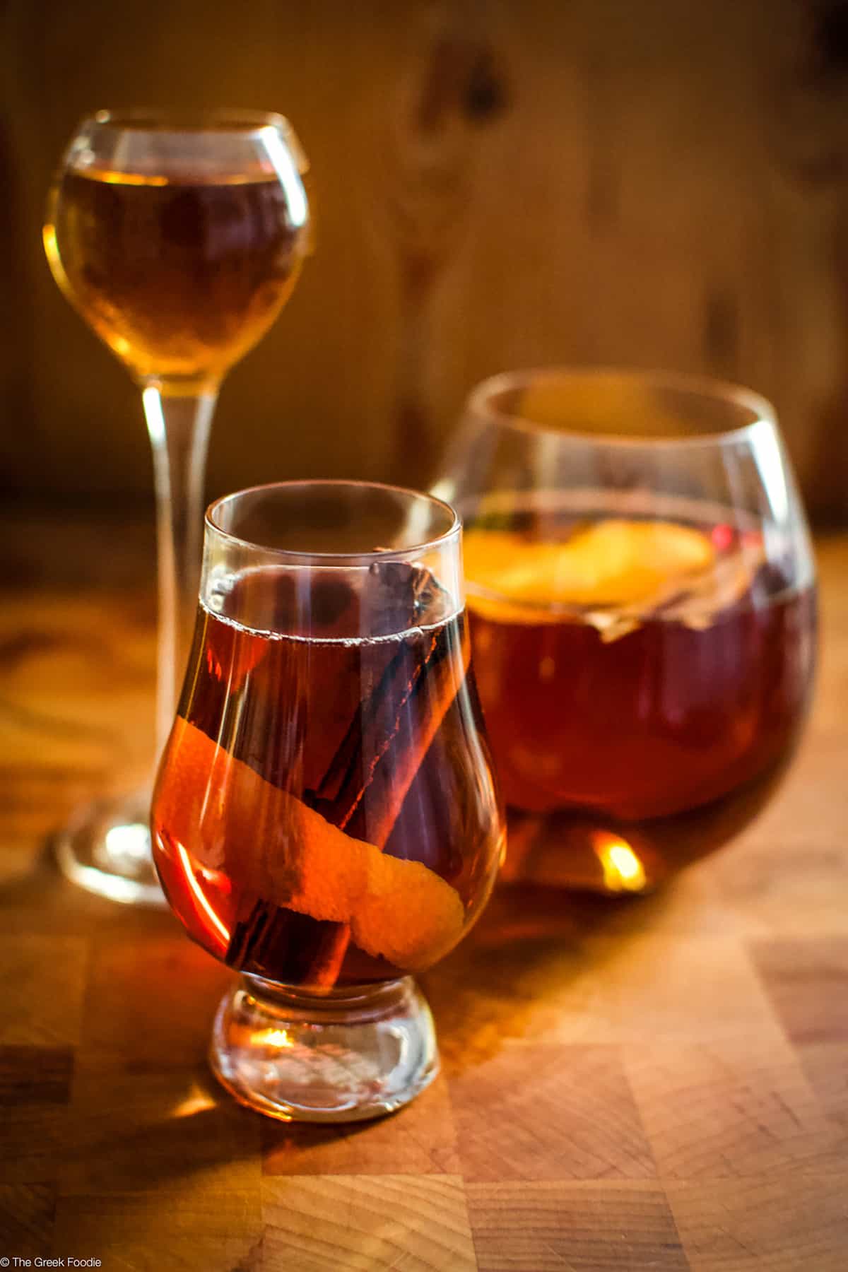 Three glasses, a tall small one, a big round and a medium glass with rakomelo, raki drink with honey in a wood environment.