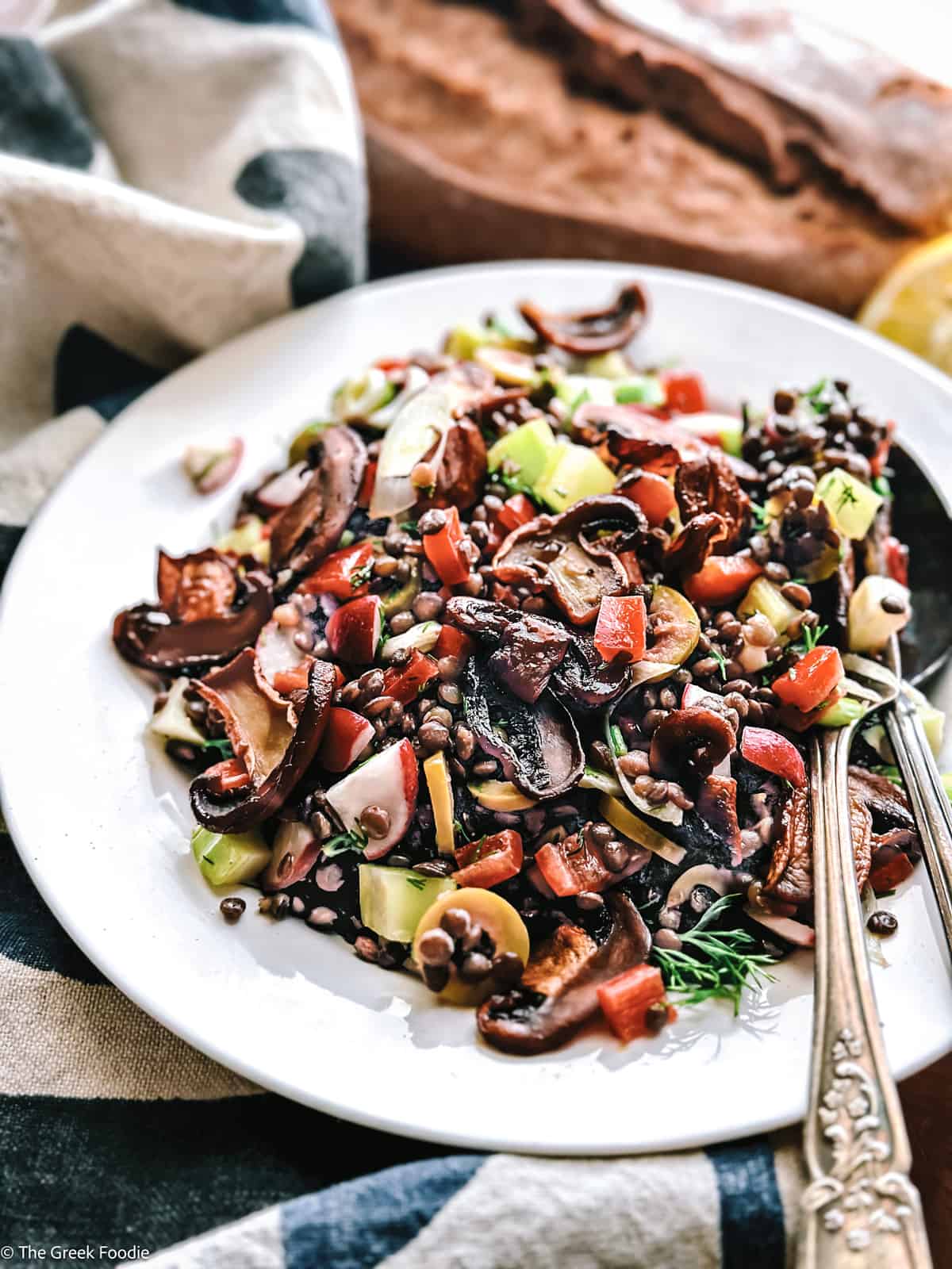 A plate with black lentils, mushrooms, scallions, peppers on a wooden table with a cloth napkin. Two halves of a lemon and a loaf of bread above it.