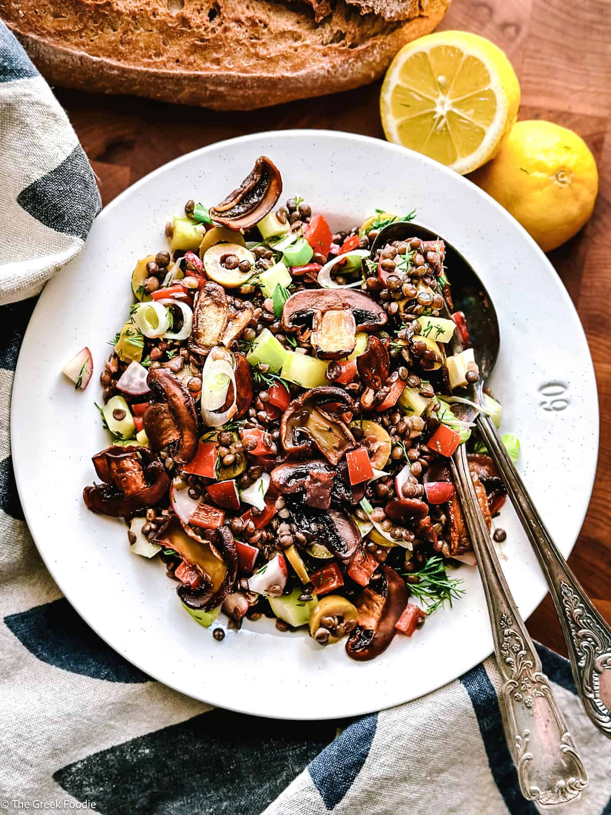 A plate with black lentils, mushrooms, scallions, peppers on a wooden table with a cloth napkin. Two halves of a lemon and a loaf of bread above it. 