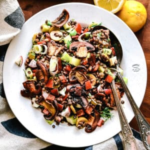 A plate with black lentils, mushrooms, scallions, peppers on a wooden table with a cloth napkin. Two halves of a lemon and a loaf of bread above it.