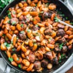 A round baking pan with roasted Gigantes plaki, carrots, sausages, pieces of graviera cheese, and chopped parsley.