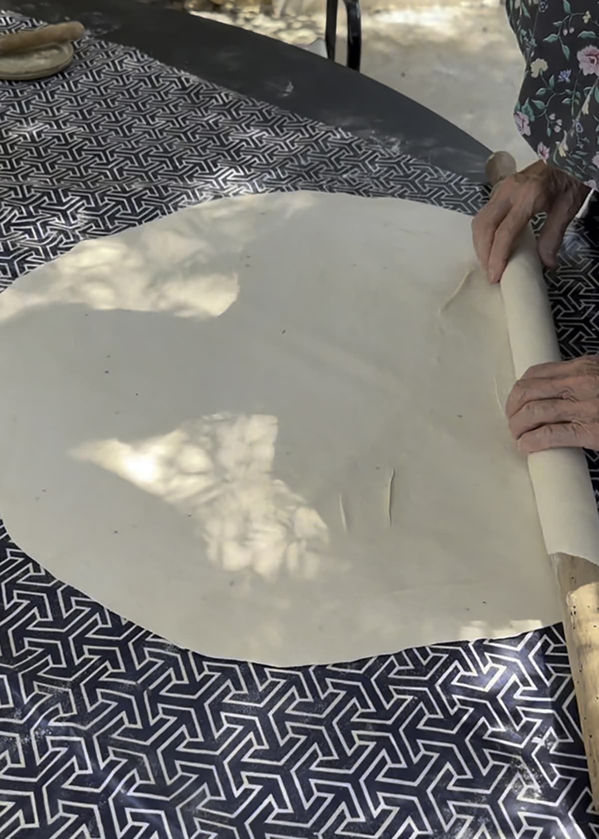 Hands of an elderly woman rolling out a piece of phyllo sheet on a table.