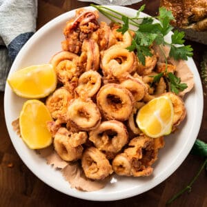 A plate with crispy fried calamari-kalamarakia with lemon wedges and parsley leaves, on a table next to a bowl with fries and a piece of bread. Overhead shot.