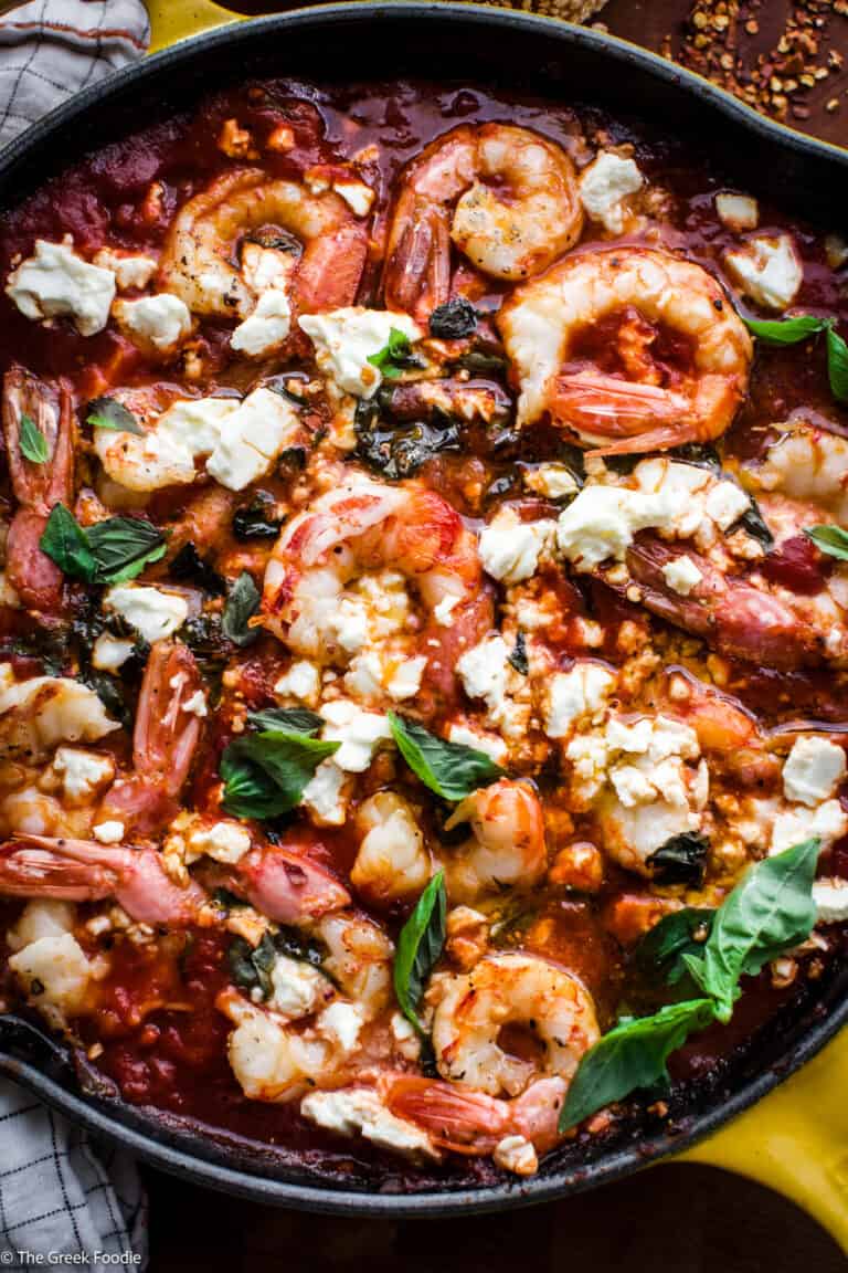 A yellow cast iron with shrimp saganaki in tomato sauce, and bread and chili flakes on a table.