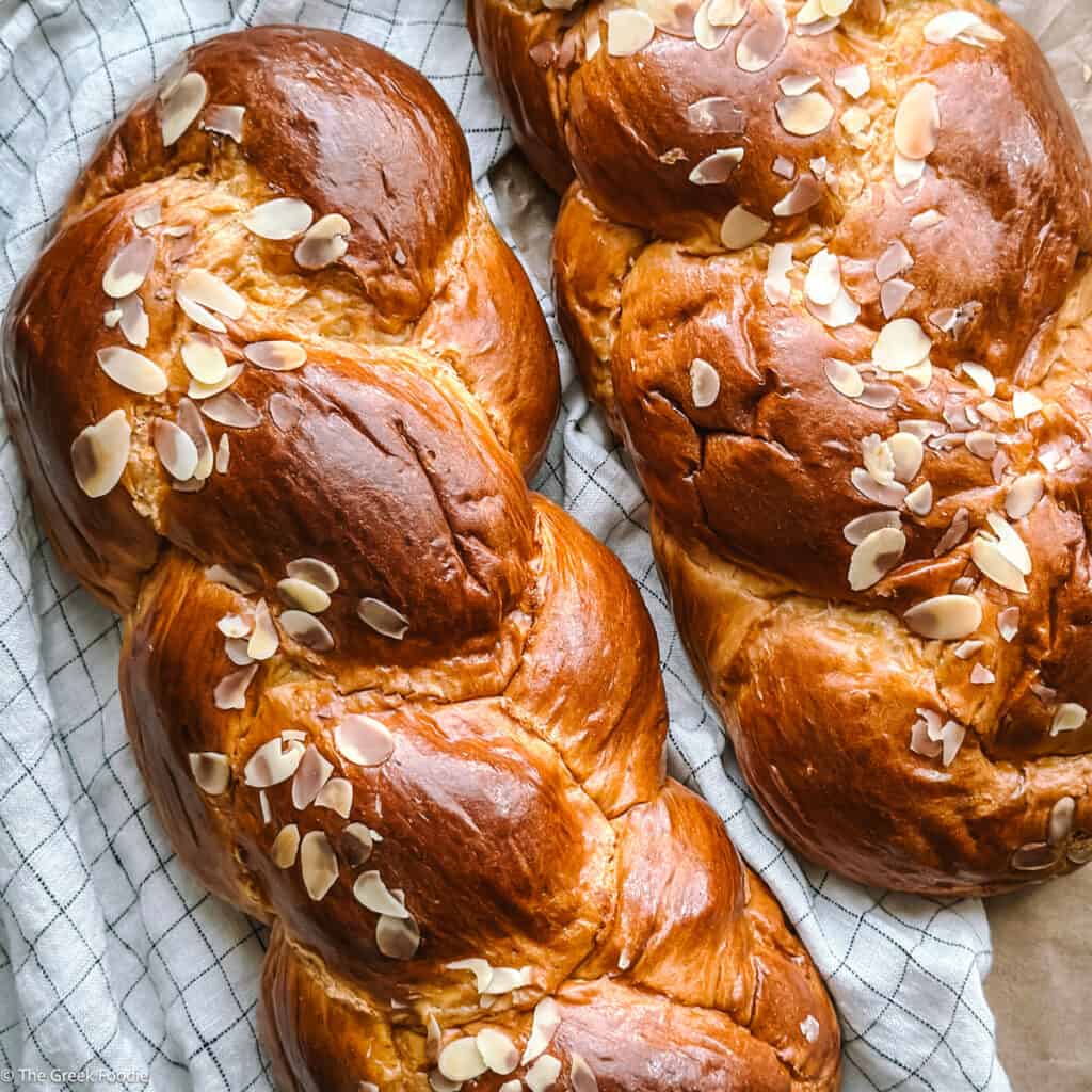 Two loaves of Tsoureki, greek brioche bread on a cloth towel on a cutting board.