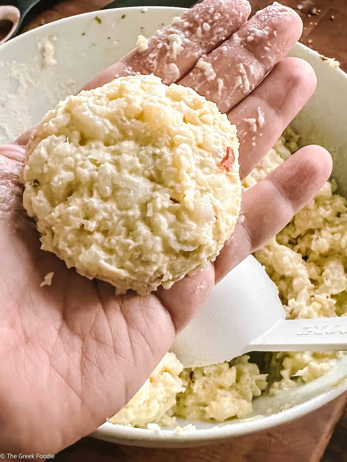 A palm of a hand with a cauliflower fritter over a bowl with cauliflower mixture.