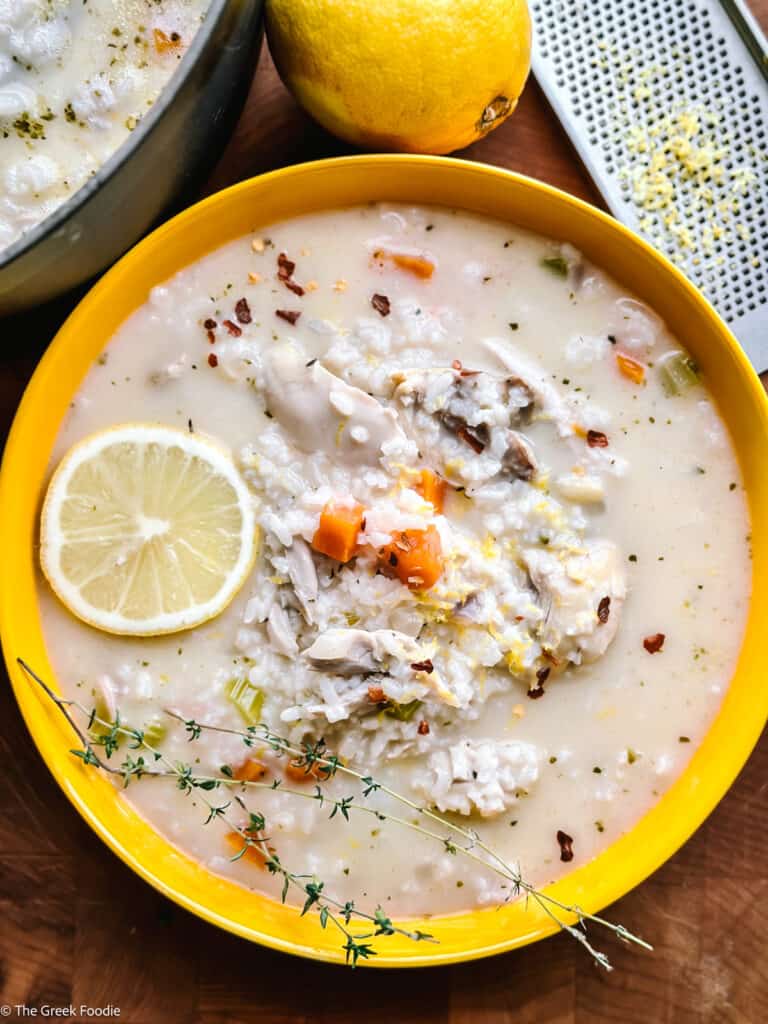 Overhead shot of avgolemono soup in a yellow bowl with a lemon slice and thyme, a whole lemon and a microplane zester alongside.