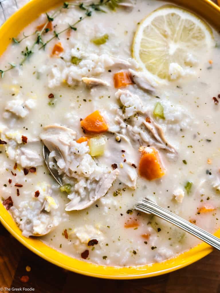 A spoon lifting shredded chicken and rice from a bowl of avgolemono soup topped with thyme and chili flakes.