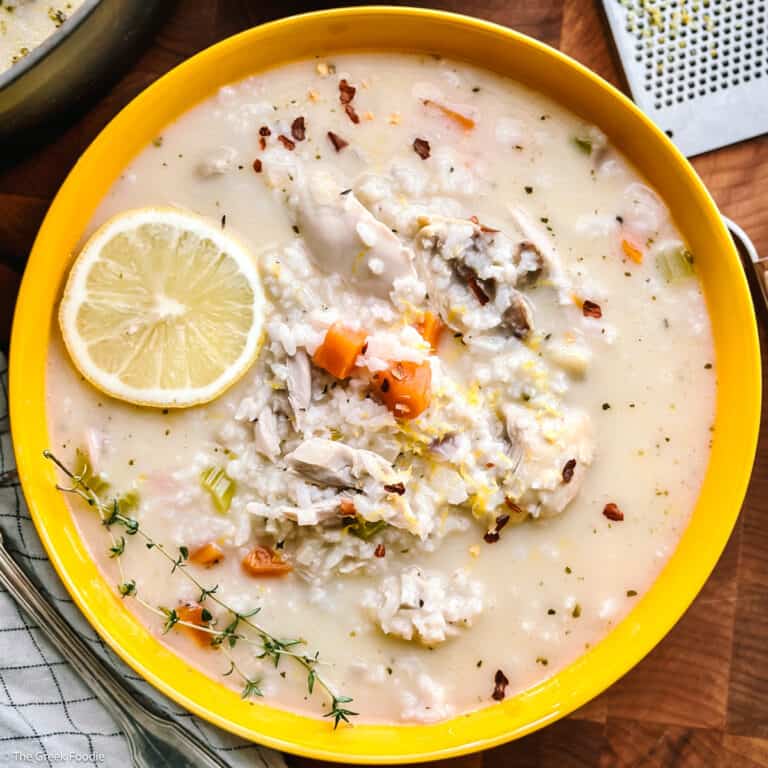 Avgolemono soup in a yellow bowl, topped with a lemon slice, fresh thyme and chili flakes, on a wooden table.