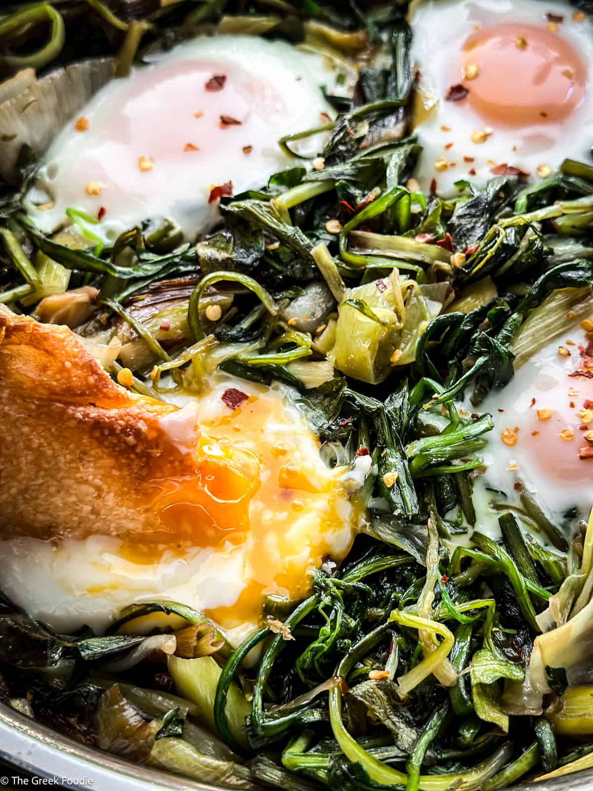 Four cooked eggs and greens. A piece of bread is breaking the egg yolk of one of the eggs.