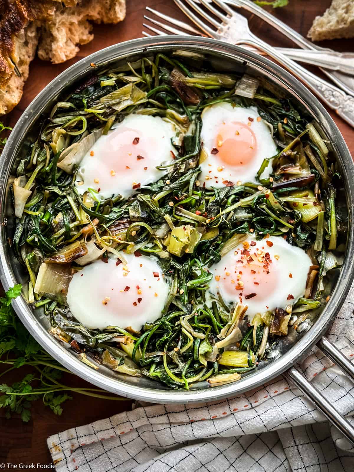 Four cooked eggs and greens in a skillet, nest to a loaf of bread, utensils and a napkin.