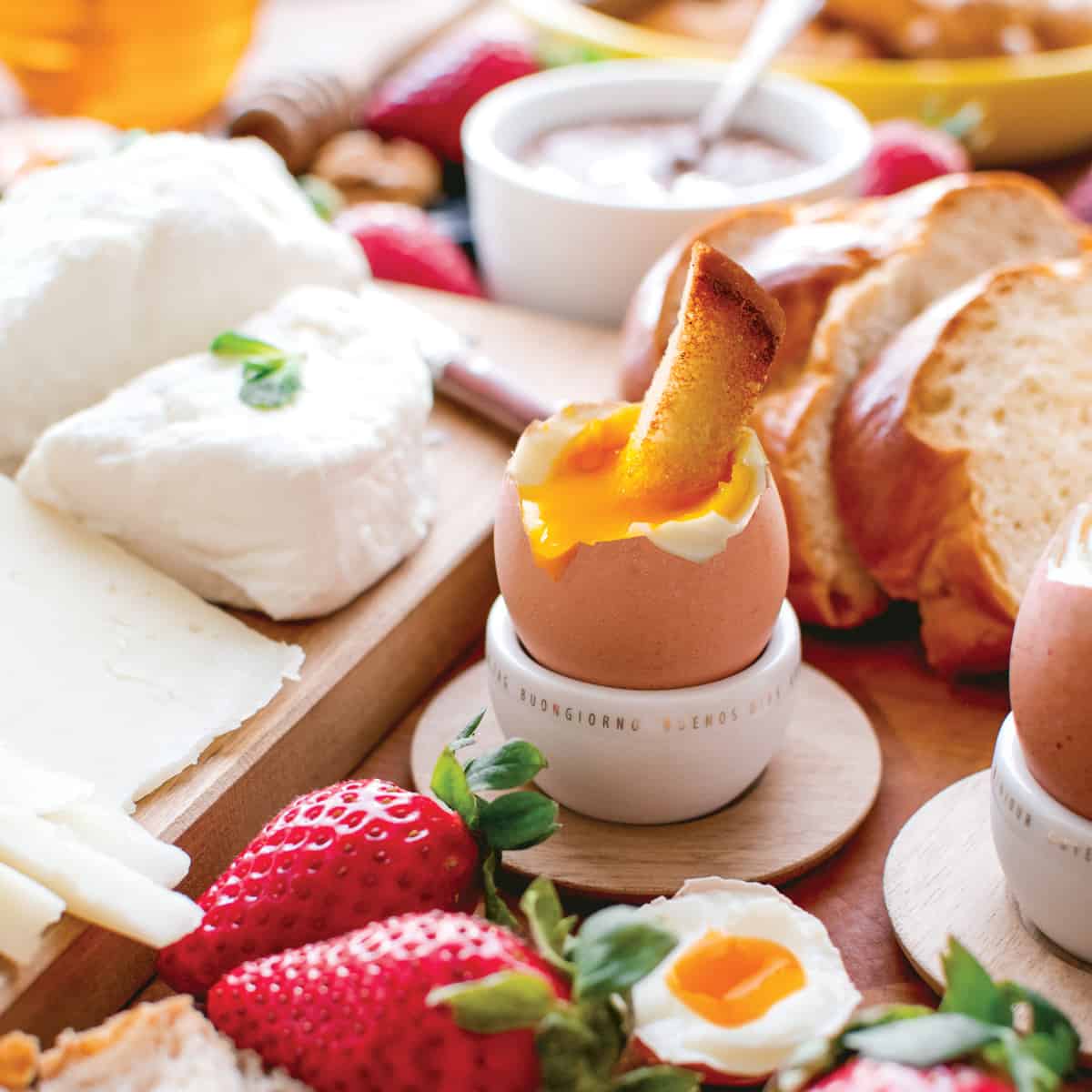 A soft boiled egg in an egg cup, broken on top with a slice of bread in the yolk, surrounded by cheese, strawberries and breads.