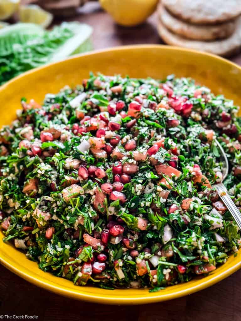Tabouli Salad-Tabbouleh in a yellow bowl on a table, at the back lettuce leaves, a lemon and pita bread.
